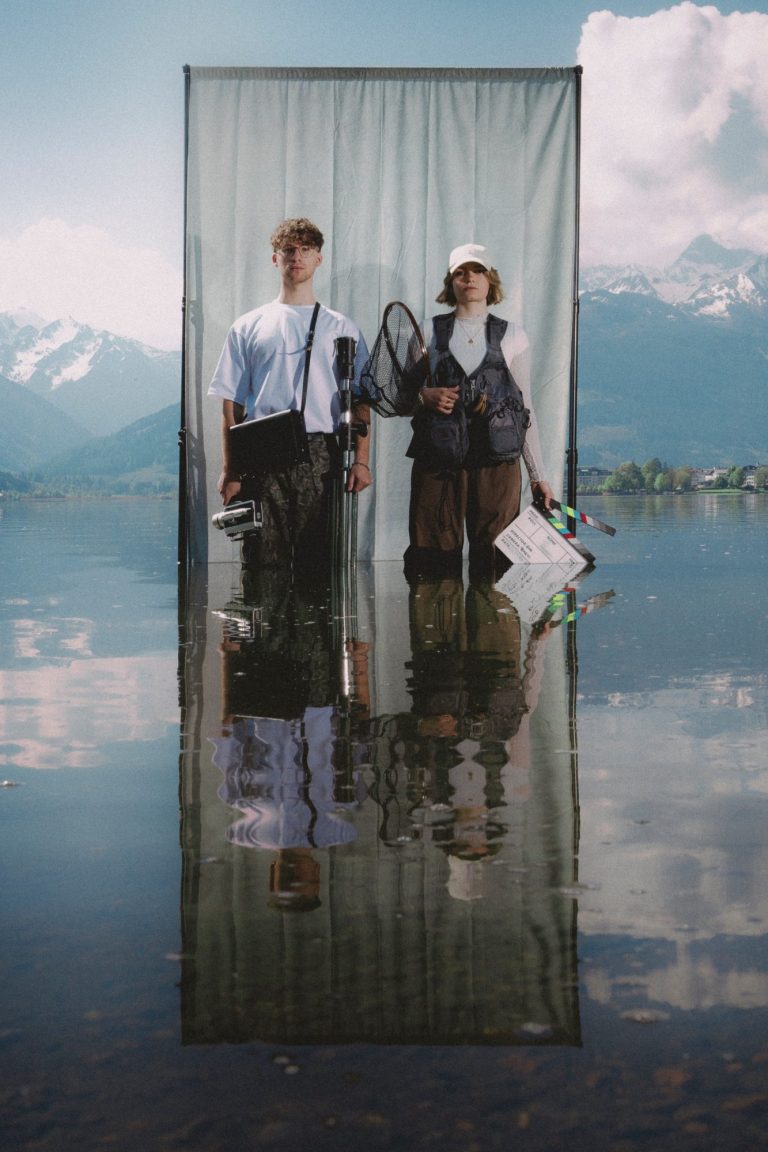 Zwei Personen stehen vor einem transparenten Vorhang am See mit Bergen im Hintergrund.